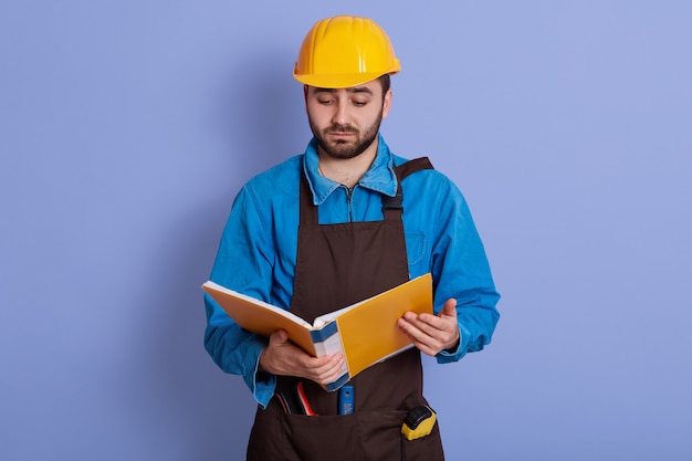 Imagen interior de un ingeniero experimentado insatisfecho molesto posando aislado sobre la pared azul en el estudio, sosteniendo una caja de papel amarilla, mirando a través de proyectos, frunciendo el ceño por la expresión facial.