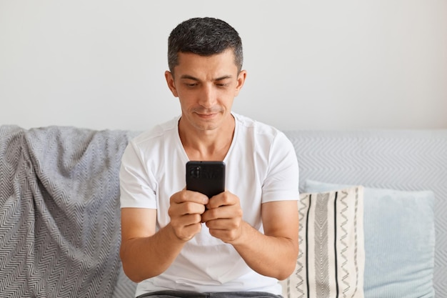 Imagen interior de un joven apuesto adulto con un pantalones blanco sentado en un sofá en casa, sosteniendo un teléfono celular, escribiendo mensajes, respondiendo a sus seguidores o escribiendo una publicación.