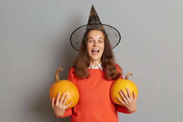 Imagen interior de una mujer caucásica sorprendida y emocionada con suéter y sombrero de bruja de pie con calabaza naranja en las manos gritando alegremente mientras celebraba Halloween posando aislado sobre fondo gris