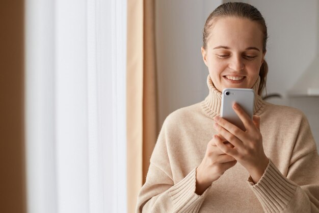 Imagen interior de una mujer optimista y encantadora con peinado de cola de caballo que usa un suéter de estilo casual beige usando un teléfono móvil para charlar con amigos o familiares, escribir masajes, sonreír.