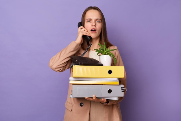 Imagen interior de una mujer sorprendida con una chaqueta beige sosteniendo muchas carpetas de documentos hablando por teléfono mirando a la cámara con grandes ojos asombrados posando aislados sobre un fondo morado