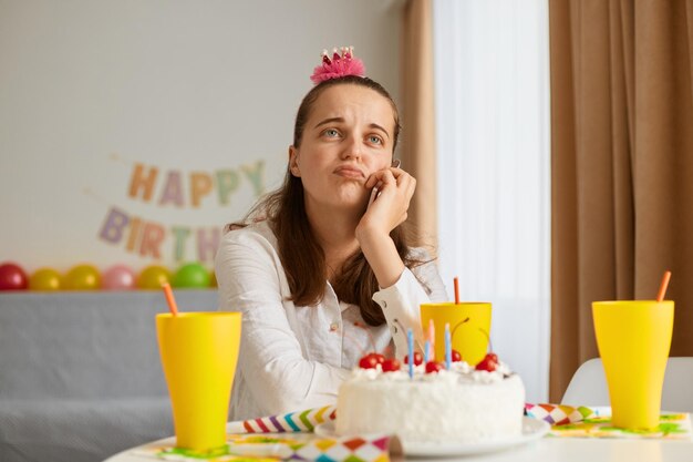 Imagen de una joven aburrida y atractiva con camisa blanca celebrando un cumpleaños sentada sola en la mesa con pastel manteniendo la mano bajo la barbilla mirando hacia otro lado soñando