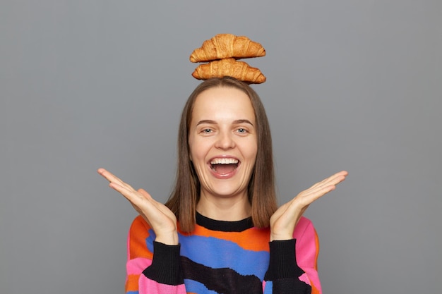 Imagen de una joven adulta caucásica extremadamente feliz y alegre que usa suéter posando aislada sobre un fondo gris sosteniendo dos croissants en la cabeza extiende las manos gritando alegremente tontamente