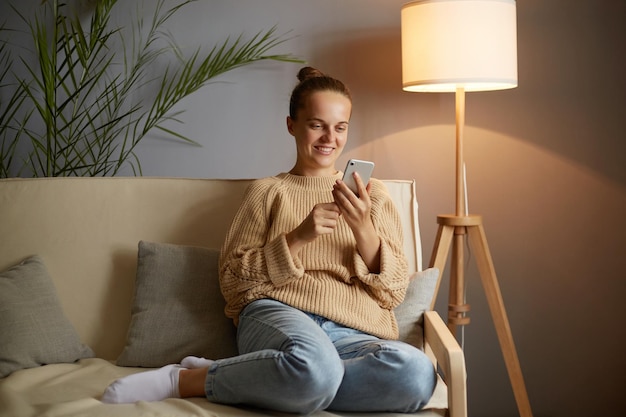 Imagen de una joven adulta sonriente y encantada con ropa informal sentada en un sofá y usando un teléfono móvil para revisar las redes sociales chateando con amigos