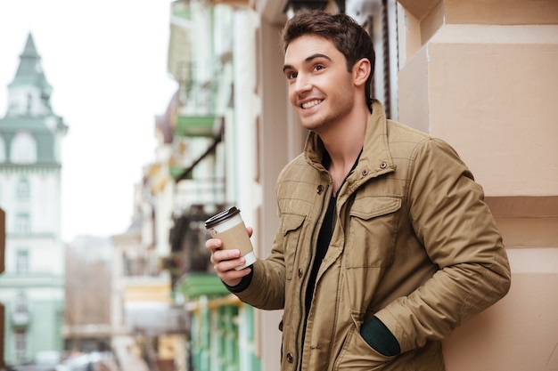 Imagen de un joven alegre caminando por la calle y mirando a un lado mientras sostiene una taza de café.