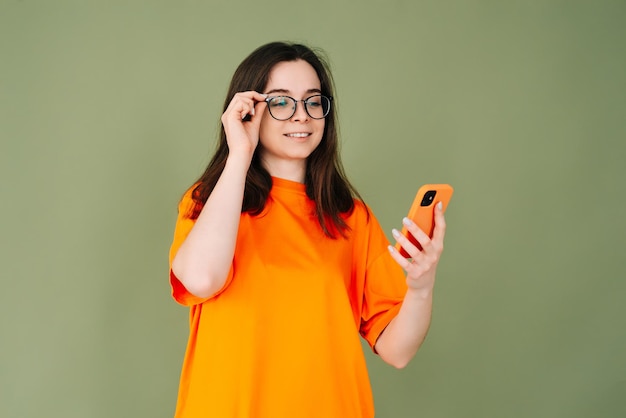 Imagen de una joven alegre con una camiseta naranja usando un smartphone moderno en un espacio verde vacío