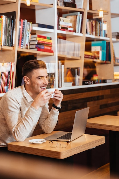 Imagen de un joven alegre sentado en la cafetería y charlando con la computadora portátil. Mirando a un lado mientras bebe café.