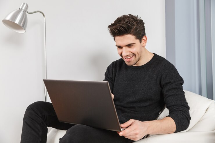Foto imagen de un joven apuesto feliz en casa en el interior con ordenador portátil.
