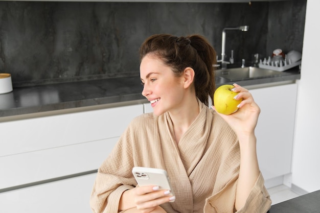 Imagen de una joven atractiva en bata de baño sentada en la cocina, come manzana verde y mira el móvil
