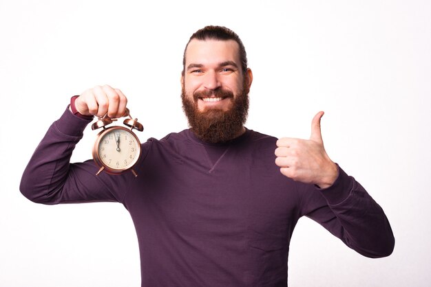 Imagen de un joven barbudo sosteniendo un reloj y mostrando un pulgar hacia arriba