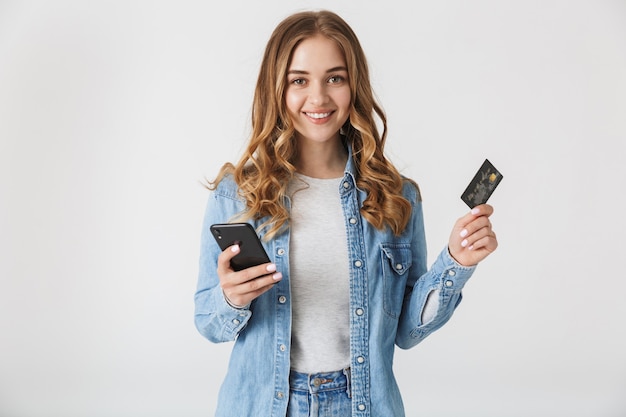 Imagen de una joven y bella mujer bonita emocionada posando aislada sobre una pared blanca mediante teléfono móvil con tarjeta de crédito.