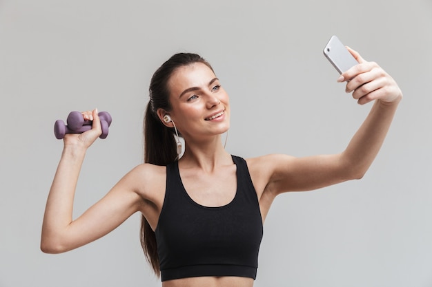 Imagen de una joven y bella mujer de fitness deporte usando teléfono móvil escuchando música aislada sobre pared gris tomar un selfie sosteniendo pesas.