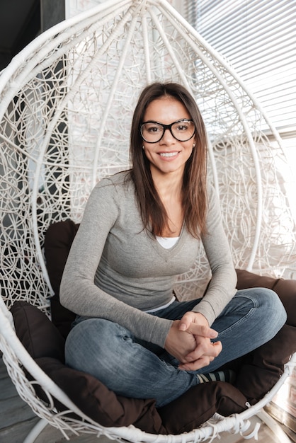 Imagen de joven bonita con anteojos sentado en una silla en el interior. Mira la cámara.