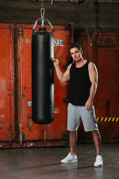 Imagen de joven boxeador fuerte de pie en un gimnasio cerca del saco de boxeo.