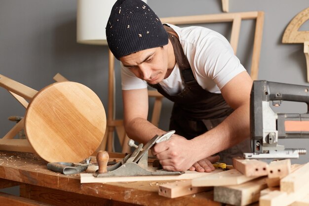 Imagen de un joven carpintero adulto con camiseta blanca, gorra negra y delantal marrón trabajando en la planificación de madera en un taller haciendo productos de madera sosteniendo un avión en las manos