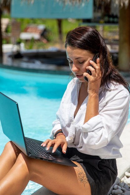 Imagen de una joven caucásica que trabaja a distancia cerrada a una piscina azul mientras usa una computadora portátil y un teléfono