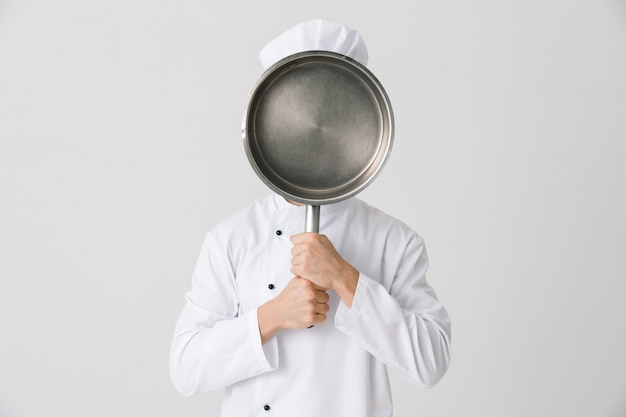 Imagen del joven chef en el interior aislado sobre fondo de pared blanca que cubre la cara con una sartén.