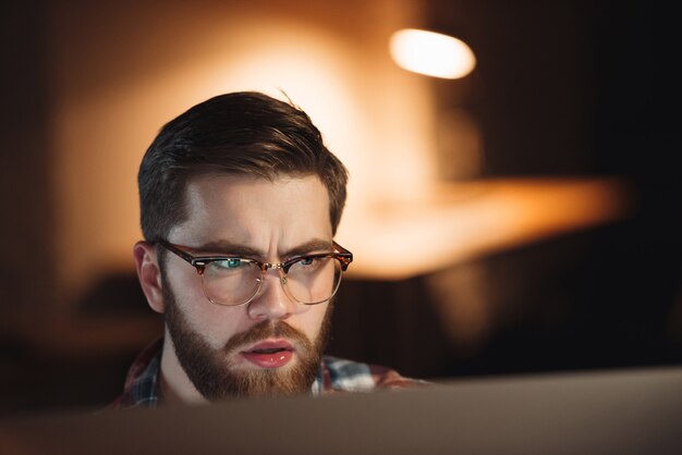 Imagen de joven diseñador web barbudo confundido vestido con camisa con estampado de jaula y gafas trabajando hasta tarde en la noche y mirando la computadora.