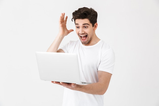 Imagen de un joven emocionado conmocionado posando aislado sobre una pared blanca usando una computadora portátil.