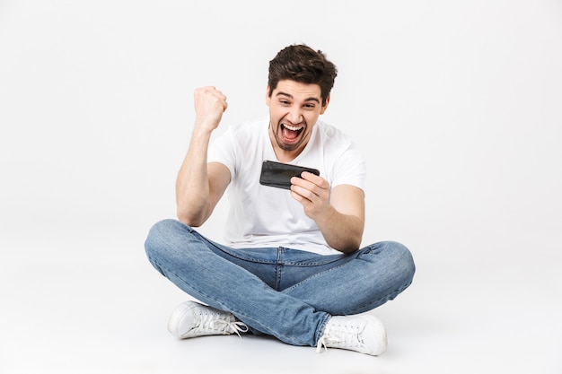 Imagen de un joven emocionado posando aislado sobre la pared blanca. Jugar juegos por teléfono móvil hace el gesto de ganador.