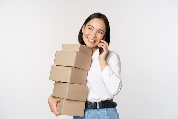 Imagen de una joven empresaria asiática que responde una llamada telefónica mientras lleva cajas para entregarlas posando sobre un fondo blanco