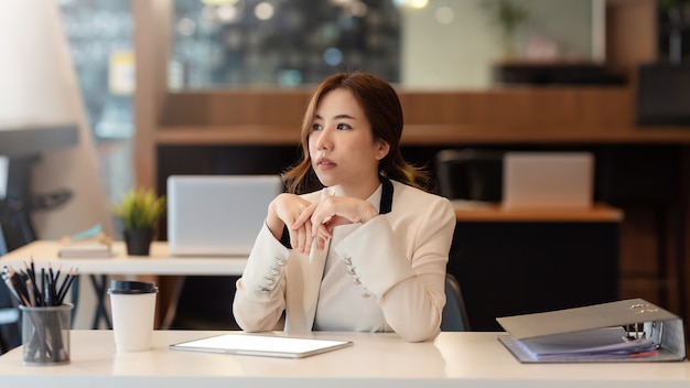 Imagen de una joven empresaria asiática sentada y meditando ideas para trabajar usando gráficos en la oficina.