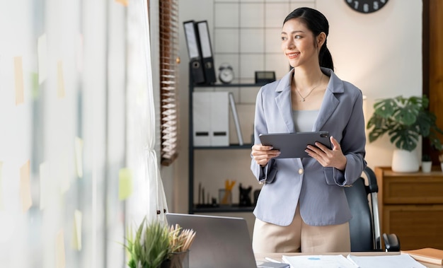 Imagen de una joven empresaria asiática trabajadora de traje sonriendo y sosteniendo una tableta digital de pie en el escritorio de la oficina