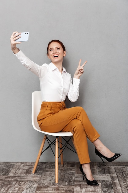 Imagen de la joven empresaria pelirroja de 20 años en ropa formal con teléfono móvil mientras está sentado en una silla en la oficina aislada sobre gris