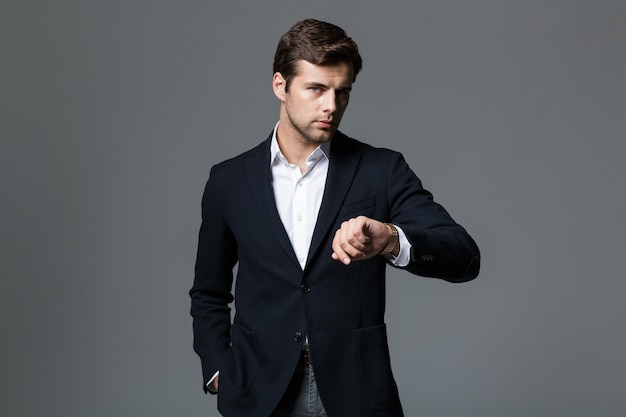 Imagen del joven empresario de 30 años en chaqueta negra mirando el reloj de pulsera, aislado sobre pared gris