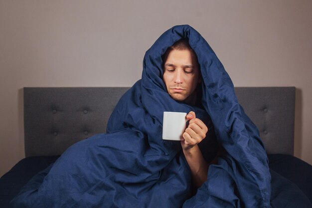 La imagen de un joven está sentado en una cama y cubierto con una manta azul oscuro. Está sosteniendo una taza de té blanca. El tipo está mirando a la cámara. Es un concepto serio de coronavirus y frío.