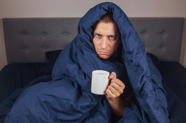 La imagen de un joven está sentado en una cama y cubierto con una manta azul oscuro. Está sosteniendo una taza de té blanca. El tipo está mirando a la cámara. Es un concepto serio de coronavirus y frío.