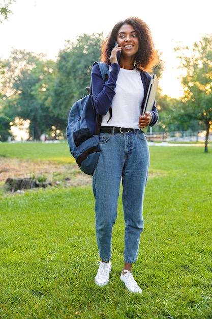 Imagen de la joven estudiante africana hermosa caminando en el parque con portátil hablando por teléfono móvil.