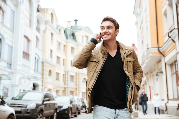 Imagen de un joven feliz caminando por la calle y mirando a un lado mientras habla por su teléfono.