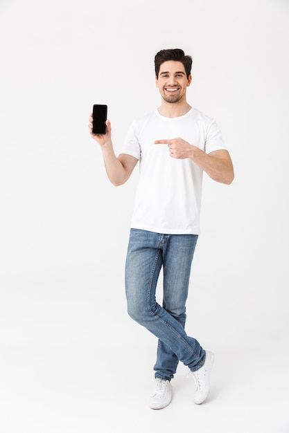 Imagen de un joven feliz emocionado posando aislado sobre una pared blanca mediante teléfono móvil que muestra la pantalla.