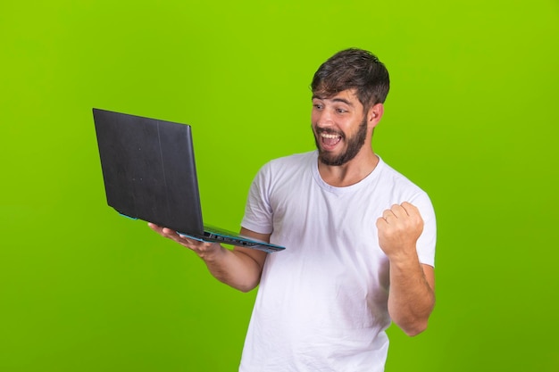 La imagen de un joven feliz y emocionado posando aislado sobre una pared verde usando una computadora portátil hace un gesto de ganador