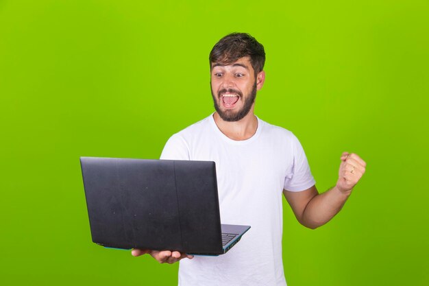 La imagen de un joven feliz y emocionado posando aislado sobre una pared verde usando una computadora portátil hace un gesto de ganador