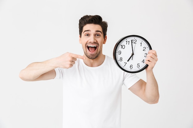 Imagen del joven feliz emocionado sorprendido posando aislado sobre la pared blanca que sostiene el reloj.