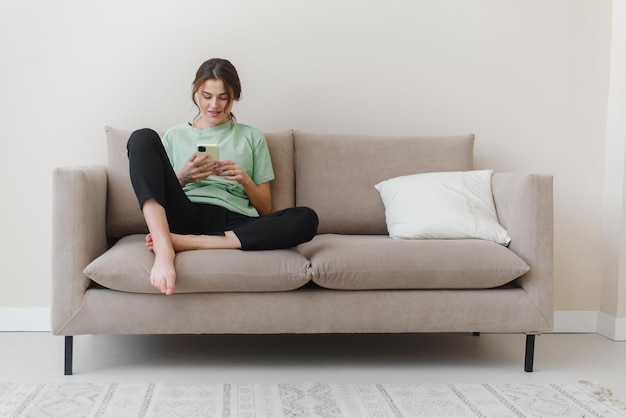 Imagen de una joven feliz y optimista sentada en el interior de su casa y comprando o en las redes sociales usando un teléfono móvil en un sofá en casa