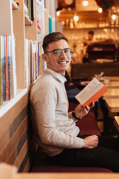 Foto imagen de joven guapo alegre sentado en la cafetería mientras lee un libro y mira a la cámara.