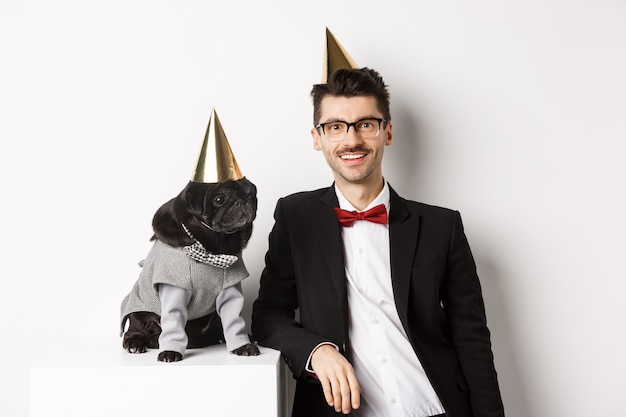 Imagen de joven guapo celebrando un cumpleaños con lindo pug negro en traje de fiesta y cono en la cabeza, de pie sobre fondo blanco.