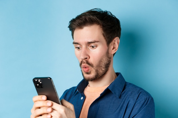 Imagen de un joven imperturbable mirando el teléfono inteligente y diciendo wow, mirando la oferta en línea, de pie sobre fondo azul.