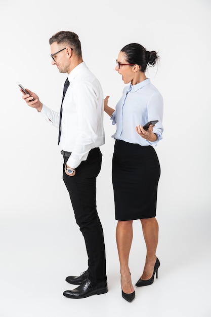 Imagen de una joven mujer gritando mirando a su colega de negocios hombre usando un teléfono móvil aislado sobre una pared blanca.