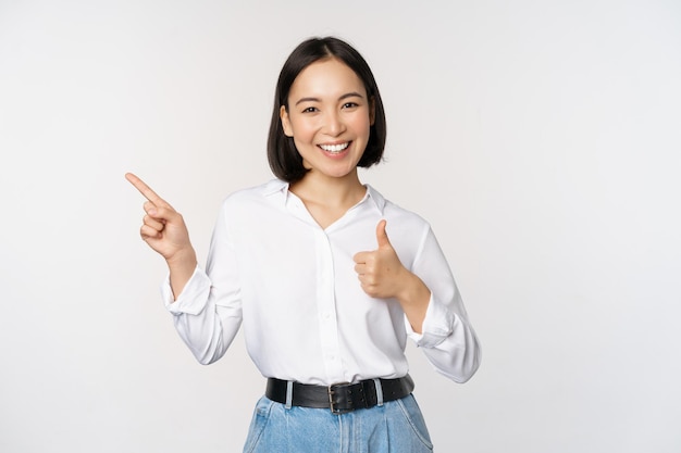 Imagen de una joven mujer de negocios asiática sonriendo mientras señala con el dedo a la izquierda y muestra el pulgar hacia arriba recomendando elogios del producto de pie sobre fondo blanco