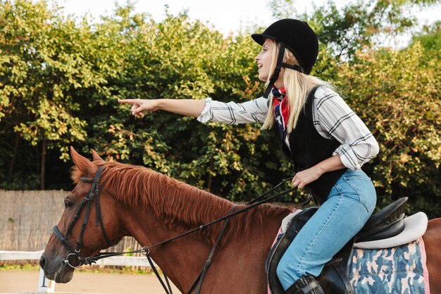 Foto imagen de joven mujer rubia con sombrero dedo acusador mientras monta a caballo en el patio en el campo