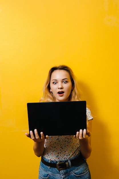 Imagen de una joven mujer sosteniendo una computadora portátil y feliz sobre pared amarilla