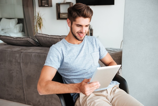 Imagen del joven nupcial vestido con camiseta gris sentado en una silla y sosteniendo la tableta