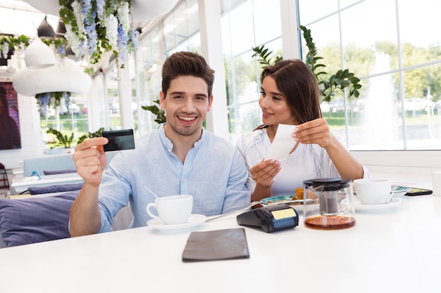 Imagen de una joven pareja amorosa feliz sentada en la cafetería con tarjeta de crédito y cheque.