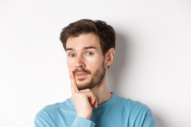 Imagen de un joven pensativo con barba, haciendo una elección, tocando el labio y mirando pensativo a la cámara, decidiendo algo, fondo blanco.