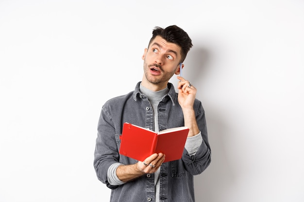 Imagen de un joven pensativo escribiendo ideas en el planificador, mirando pensativo el logo y rascándose la oreja con un bolígrafo, sosteniendo el diario en las manos, fondo blanco.