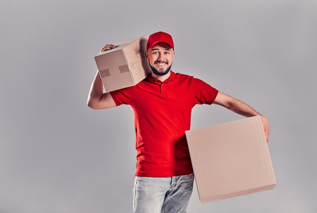 Imagen de un joven repartidor feliz con gorra roja de pie con buzones de paquetería aislados sobre fondo gris.